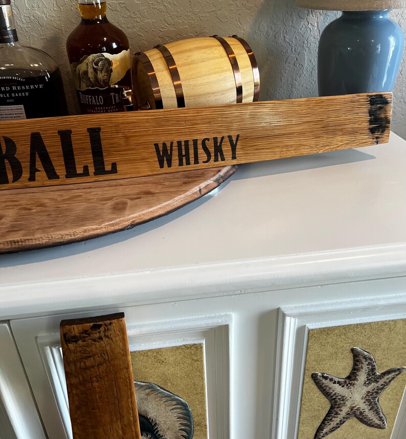 Puede incluir: Un letrero de madera con la palabra "WHISKY" en letras negras. El letrero est&aacute; hecho de un trozo de barril y tiene un aspecto r&uacute;stico. Otros elementos en la imagen incluyen un peque&ntilde;o barril de madera y botellas de whisky.