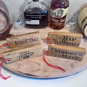 May include: Wooden bourbon barrel stave tags with the names "Buffalo Trace", "Kentucky Bourbon Trail", "Old Rip Van Winkle", and "Maker's Mark" printed on them. Each tag has a red string attached.