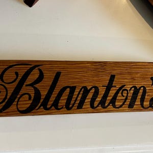 Puede incluir: Un letrero rectangular de madera con el nombre "Blanton's" en escritura negra. El letrero est&aacute; hecho de madera marr&oacute;n claro con vetas visibles y es probablemente un art&iacute;culo decorativo.