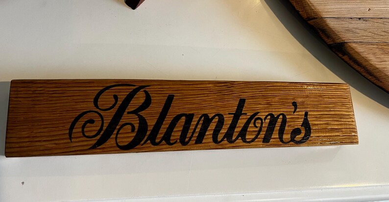 Puede incluir: Un letrero rectangular de madera con el nombre "Blanton's" en escritura negra. El letrero est&aacute; hecho de madera marr&oacute;n claro con vetas visibles y es probablemente un art&iacute;culo decorativo.