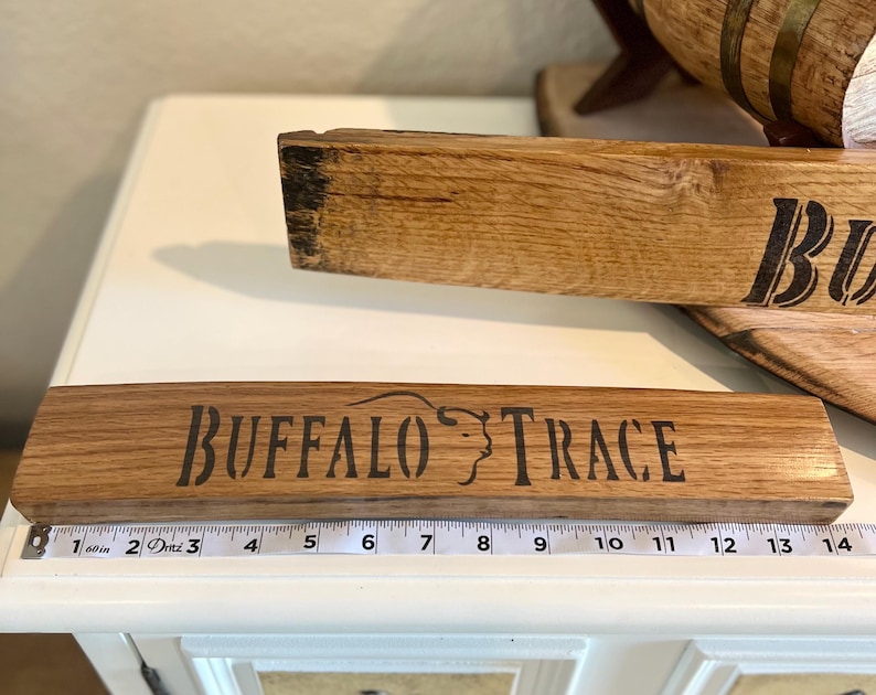 Puede incluir: Dos tablas de madera con las palabras "BUFFALO TRACE" y "BU" en letras negras. La tabla inferior mide aproximadamente 35,5 cm de largo. La madera tiene una veta natural y es de color marr&oacute;n claro.