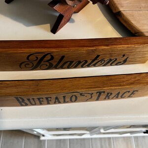 Puede incluir: Dos letreros de madera con bordes angulados. El letrero superior dice "Blanton's" en escritura negra. El letrero inferior dice "BUFFALO TRACE" en letras may&uacute;sculas negras. Ambos letreros son de madera marr&oacute;n.