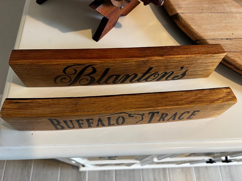 Puede incluir: Dos letreros de madera con bordes angulados. El letrero superior dice "Blanton's" en escritura negra. El letrero inferior dice "BUFFALO TRACE" en letras may&uacute;sculas negras. Ambos letreros son de madera marr&oacute;n.