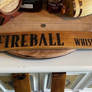 Puede incluir: Un letrero de madera curvado con las palabras "FIREBALL WHISKY" en letras negras. El letrero es de madera de color marr&oacute;n claro. El fondo incluye una barrica de madera y botellas de licor.