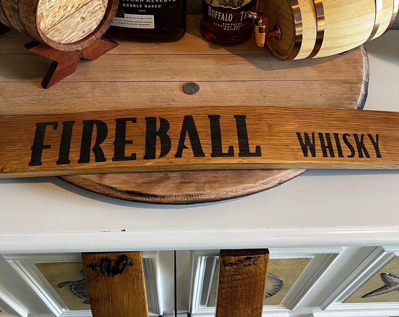 Puede incluir: Un letrero de madera curvado con las palabras "FIREBALL WHISKY" en letras negras. El letrero es de madera de color marr&oacute;n claro. El fondo incluye una barrica de madera y botellas de licor.