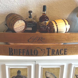 Op de afbeelding: Houten duigenborden met de tekst "Eagle Rare" en "Buffalo Trace". De afbeelding toont ook twee kleine houten vaten en twee flessen bourbon. De items worden weergegeven op een witte kast.