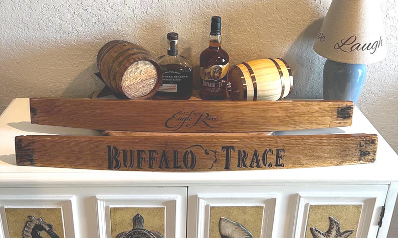 Op de afbeelding: Houten duigenborden met de tekst "Eagle Rare" en "Buffalo Trace". De afbeelding toont ook twee kleine houten vaten en twee flessen bourbon. De items worden weergegeven op een witte kast.