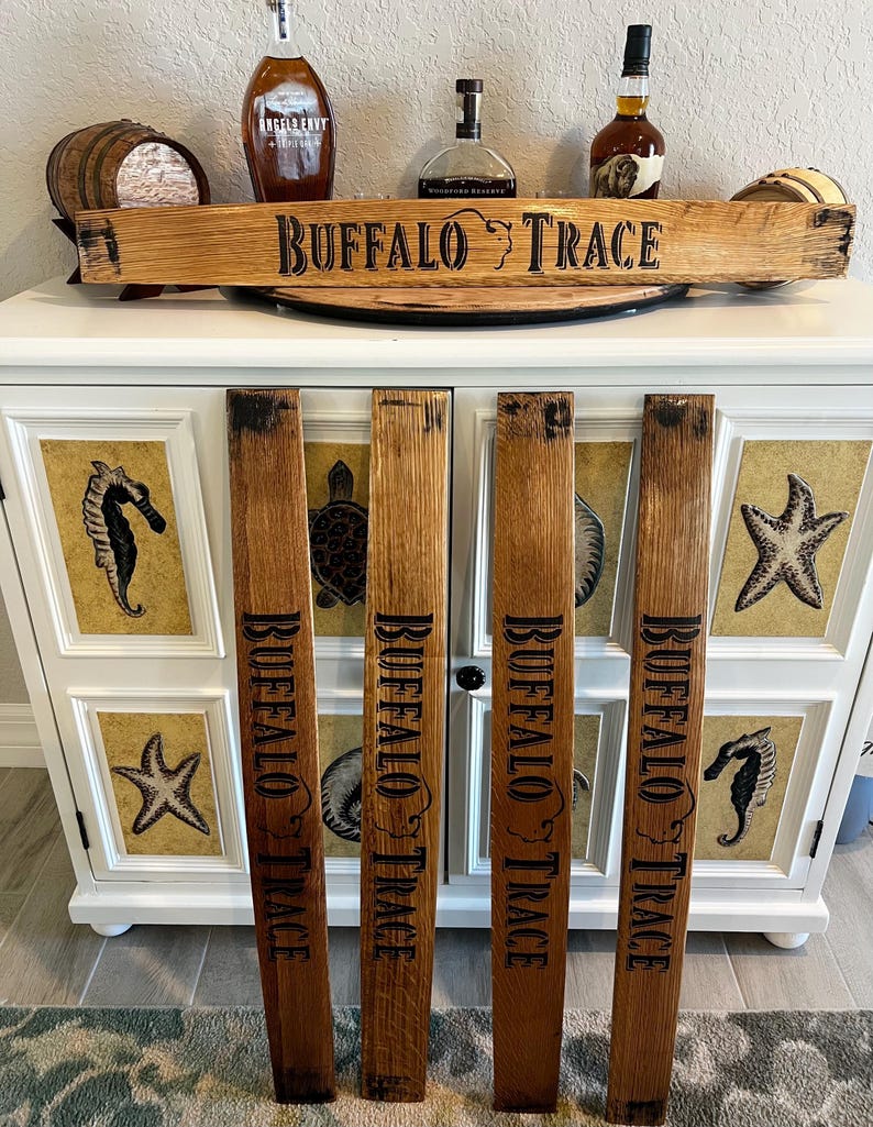 May include: Wooden sign and staves with "BUFFALO TRACE" text. The sign is made from a barrel stave and the staves are vertically placed. The sign and staves are brown with black lettering. The background includes liquor bottles and a white cabinet.