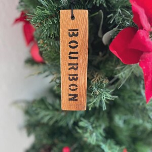 Puede incluir: Adorno de &aacute;rbol de Navidad de madera con la palabra "BOURBON" impresa en negro. El adorno rectangular cuelga de una cuerda negra. El fondo presenta un &aacute;rbol de Navidad verde con lazos rojos.