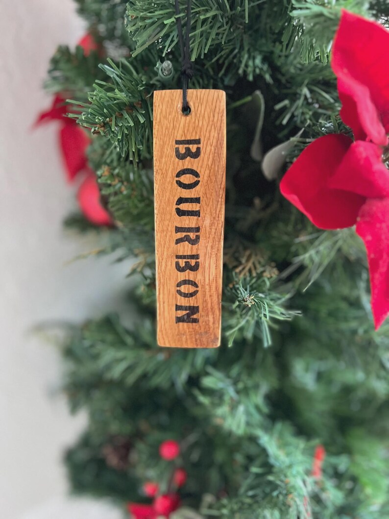 Puede incluir: Adorno de &aacute;rbol de Navidad de madera con la palabra "BOURBON" impresa en negro. El adorno rectangular cuelga de una cuerda negra. El fondo presenta un &aacute;rbol de Navidad verde con lazos rojos.