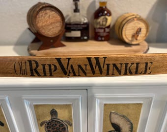May include: A wooden barrel stave with the words "Old Rip Van Winkle" in black lettering. The stave is in front of a white cabinet with decorative panels. Behind the stave are small wooden barrels and bottles.