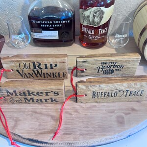 May include: Wooden bourbon barrel stave tags with the names "Old Rip Van Winkle", "Maker's Mark", "Kentucky Bourbon Trail", and "Buffalo Trace" printed on them. Each tag has a red string attached. Bourbon bottles and glasses are in the background.