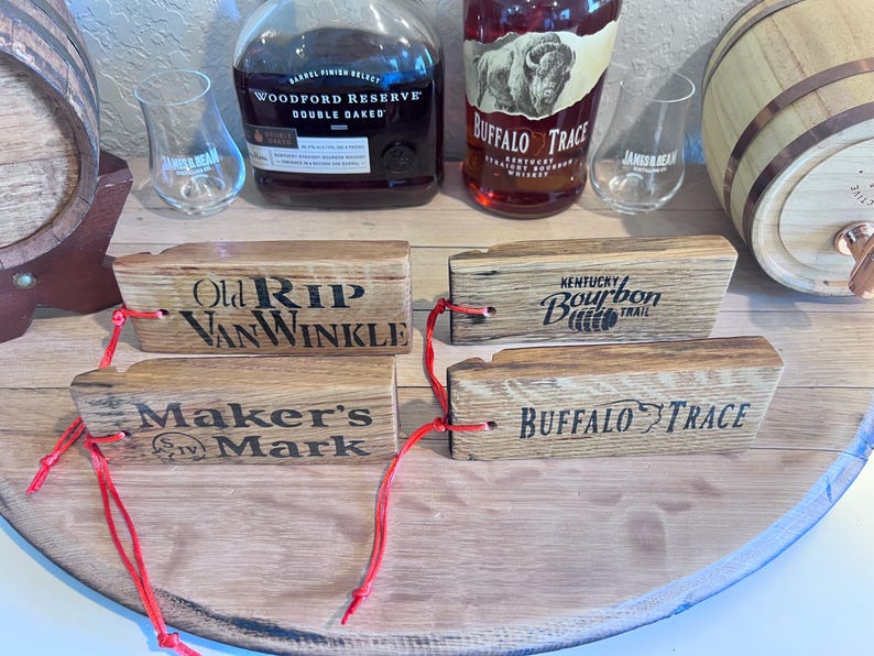 May include: Wooden bourbon barrel stave tags with the names "Old Rip Van Winkle", "Maker's Mark", "Kentucky Bourbon Trail", and "Buffalo Trace" printed on them. Each tag has a red string attached. Bourbon bottles and glasses are in the background.