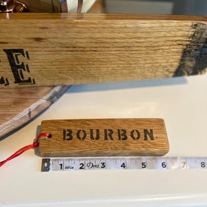 Puede incluir: Una etiqueta rectangular de madera con la palabra "BOURBON" estampada en negro. La etiqueta est&aacute; sujeta a una cuerda roja. La etiqueta mide aproximadamente 13 cm de largo. La etiqueta es de madera.