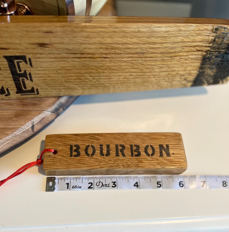 Puede incluir: Una etiqueta rectangular de madera con la palabra "BOURBON" estampada en negro. La etiqueta est&aacute; sujeta a una cuerda roja. La etiqueta mide aproximadamente 13 cm de largo. La etiqueta es de madera.