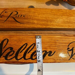 May include: Two wooden signs with black cursive text, one reading "Le Rare" and the other "Vellor". A measuring tape shows the sign is approximately 3 inches wide. The signs are made of light brown wood.