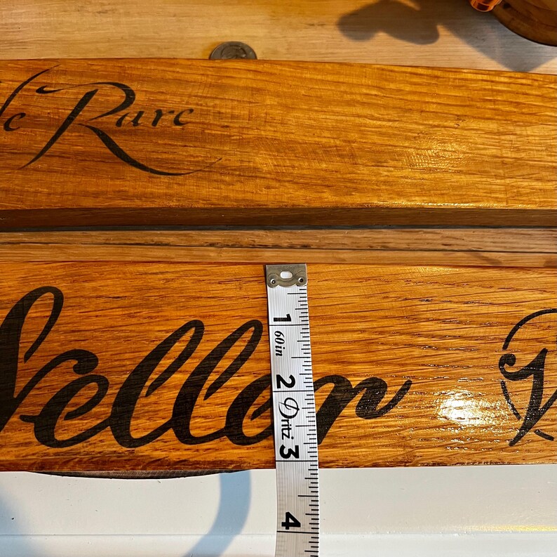 May include: Two wooden signs with black cursive text, one reading "Le Rare" and the other "Vellor". A measuring tape shows the sign is approximately 3 inches wide. The signs are made of light brown wood.