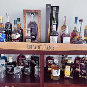 May include: A dark wooden bar cart displaying various bottles of liquor, including bourbon and whiskey. The bottles are of different shapes and sizes, with a wooden sign that reads "Buffalo Trace". Several clear glasses are also visible.