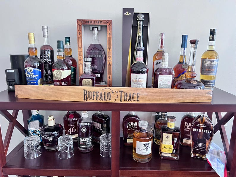 May include: A dark wooden bar cart displaying various bottles of liquor, including bourbon and whiskey. The bottles are of different shapes and sizes, with a wooden sign that reads "Buffalo Trace". Several clear glasses are also visible.