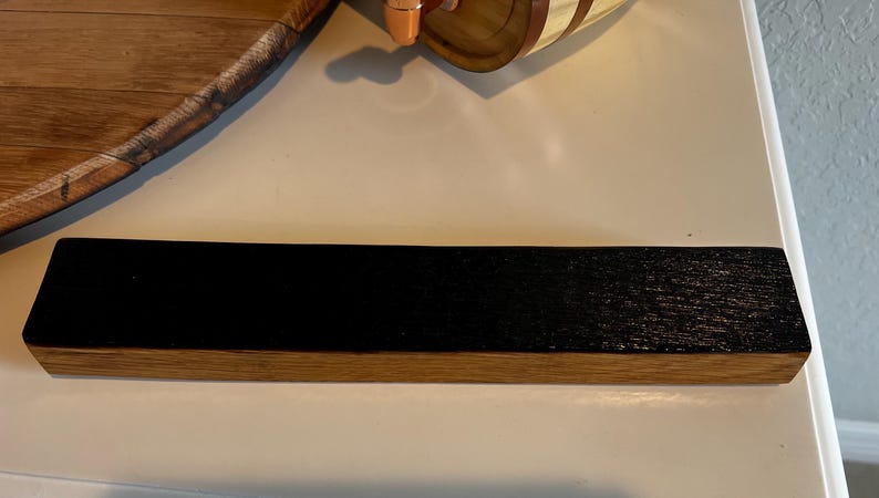 Puede incluir: Un objeto rectangular de madera con una parte superior te&ntilde;ida de negro oscuro y una base de color madera natural. El art&iacute;culo parece ser una peque&ntilde;a estanter&iacute;a o pieza de exhibici&oacute;n, mostrando un contraste entre los tonos de madera oscuros y claros.