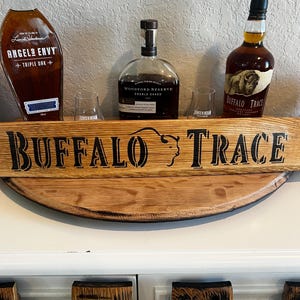 May include: A wooden shelf with three bottles of whiskey and two glasses. The shelf has the words "BUFFALO TRACE" in large black letters. The bottles are labeled "ANGELS ENVY", "WOODFORD RESERVE", and "BUFFALO TRACE".