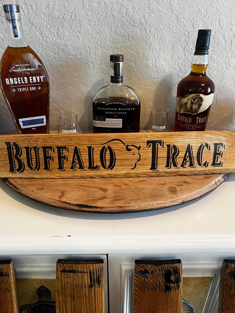 May include: A wooden shelf with three bottles of whiskey and two glasses. The shelf has the words "BUFFALO TRACE" in large black letters. The bottles are labeled "ANGELS ENVY", "WOODFORD RESERVE", and "BUFFALO TRACE".