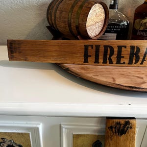 Puede incluir: Un letrero de madera con la palabra "FIREBALL" en letras negras. Un peque&ntilde;o barril de madera est&aacute; detr&aacute;s del letrero. El letrero y el barril est&aacute;n sobre una superficie de madera. Dos botellas de licor est&aacute;n en el fondo.