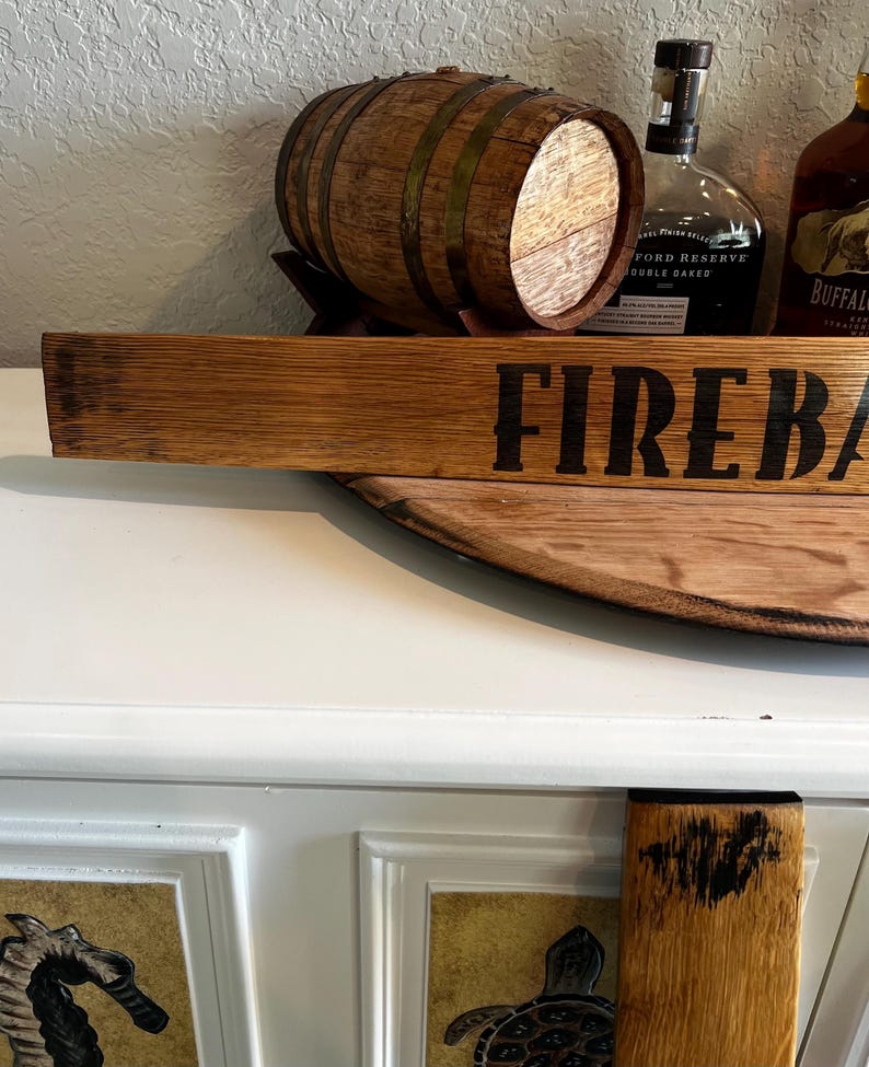 Puede incluir: Un letrero de madera con la palabra "FIREBALL" en letras negras. Un peque&ntilde;o barril de madera est&aacute; detr&aacute;s del letrero. El letrero y el barril est&aacute;n sobre una superficie de madera. Dos botellas de licor est&aacute;n en el fondo.