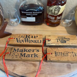 May include: Four wooden bourbon barrel stave tags with red string. The tags are branded with the names "Old Rip Van Winkle", "Maker's Mark", "Kentucky Bourbon Trail", and "Buffalo Trace". Bottles of bourbon and a barrel are in the background.