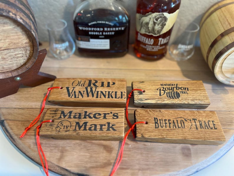 May include: Four wooden bourbon barrel stave tags with red string. The tags are branded with the names "Old Rip Van Winkle", "Maker's Mark", "Kentucky Bourbon Trail", and "Buffalo Trace". Bottles of bourbon and a barrel are in the background.