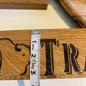 Puede incluir: Primer plano de un letrero de madera con letras negras y adornos decorativos. Una cinta m&eacute;trica muestra que el letrero mide aproximadamente 7,6 cm de ancho. El texto del letrero es parcialmente visible.
