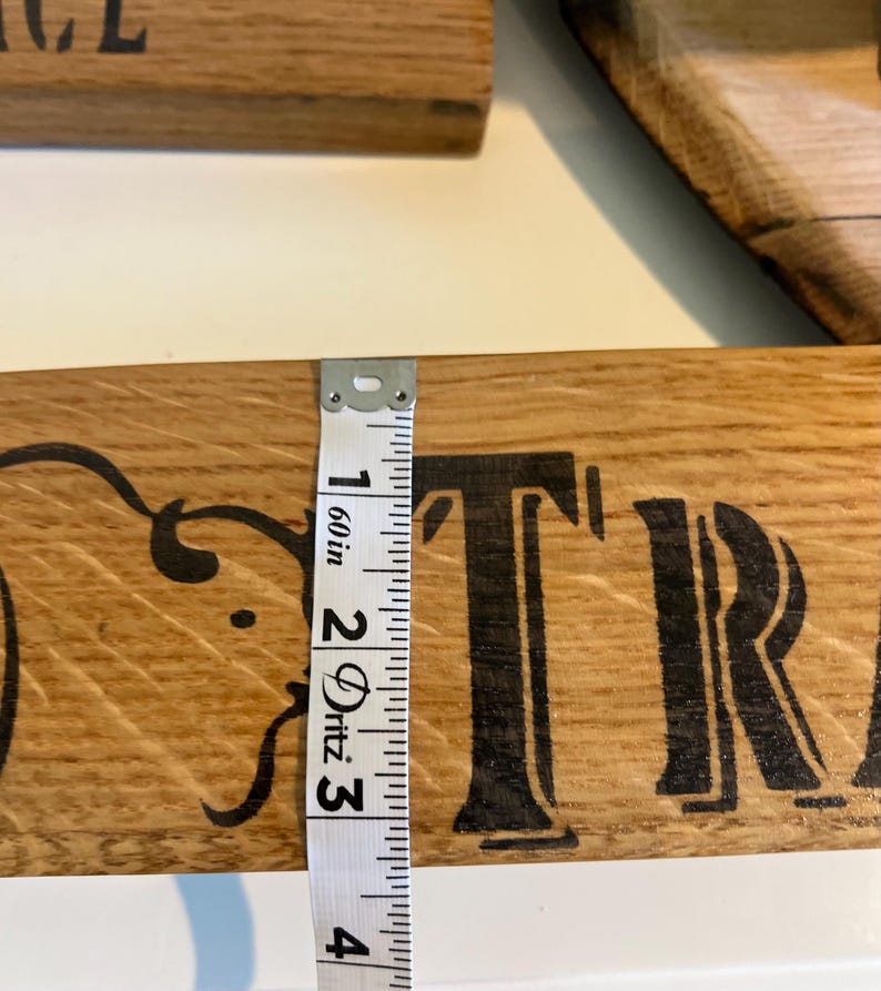Puede incluir: Primer plano de un letrero de madera con letras negras y adornos decorativos. Una cinta m&eacute;trica muestra que el letrero mide aproximadamente 7,6 cm de ancho. El texto del letrero es parcialmente visible.