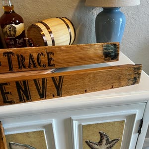 May include: Wooden planks with the words "TRACE" and "ENVY" are displayed. A bottle of Buffalo Trace bourbon, a small wooden barrel, and a blue lamp with the word "Laugh" are also visible. The cabinet below has seashell and starfish designs.