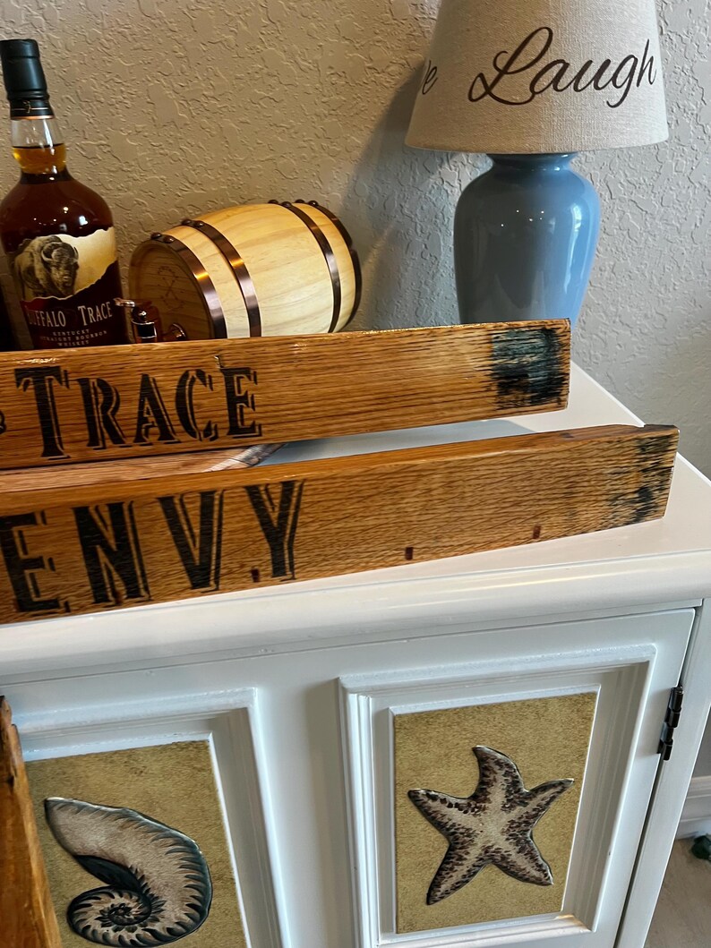 May include: Wooden planks with the words "TRACE" and "ENVY" are displayed. A bottle of Buffalo Trace bourbon, a small wooden barrel, and a blue lamp with the word "Laugh" are also visible. The cabinet below has seashell and starfish designs.