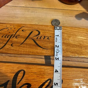 May include: A wooden sign with the words "Eagle Rare" and "Welcome" in a cursive font. A measuring tape shows the sign is approximately 5 inches long. A quarter is placed on the sign. Two bottles of whiskey are in the background.