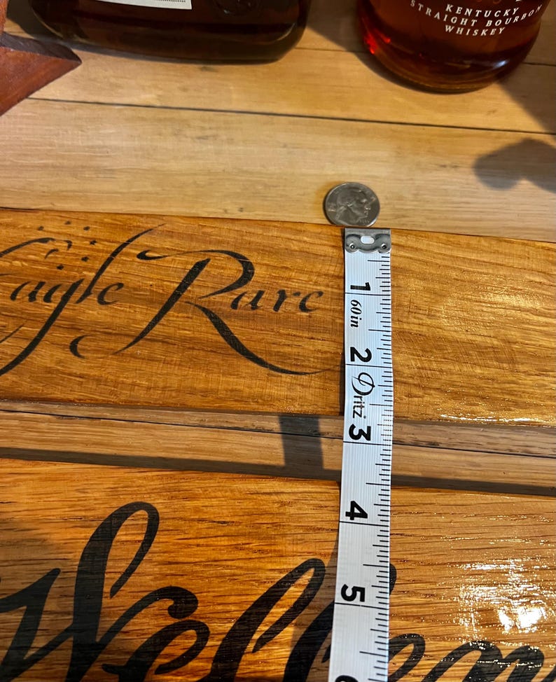 May include: A wooden sign with the words "Eagle Rare" and "Welcome" in a cursive font. A measuring tape shows the sign is approximately 5 inches long. A quarter is placed on the sign. Two bottles of whiskey are in the background.