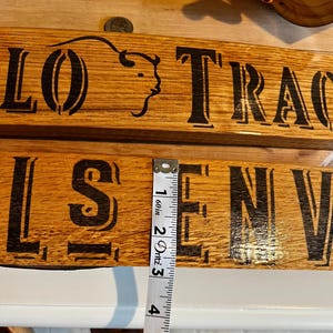 May include: Two wooden signs with black lettering and a buffalo head logo. The top sign reads "LO TRA", and the bottom sign reads "LS EN". A measuring tape shows the sign is approximately 4 inches long.