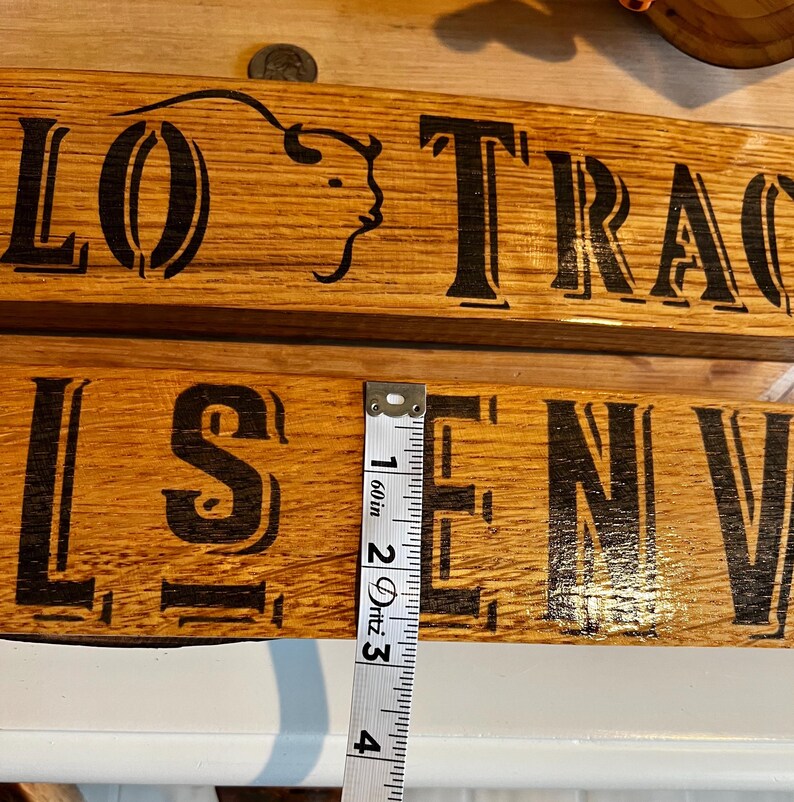 May include: Two wooden signs with black lettering and a buffalo head logo. The top sign reads "LO TRA", and the bottom sign reads "LS EN". A measuring tape shows the sign is approximately 4 inches long.