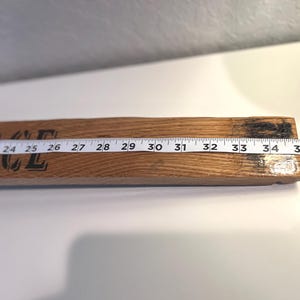 May include: A wooden sign with the word "GRACE" printed in black. A white measuring tape is laid across the sign, showing measurements from 22 to 36 inches. The wood has a natural grain and a dark, charred end.