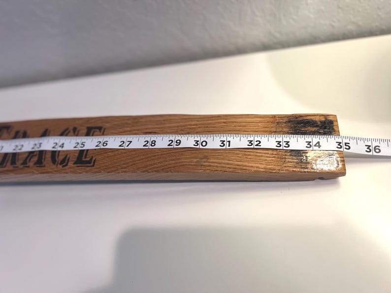 May include: A wooden sign with the word "GRACE" printed in black. A white measuring tape is laid across the sign, showing measurements from 22 to 36 inches. The wood has a natural grain and a dark, charred end.