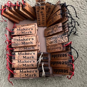 May include: A collection of wooden tags with various brand names and logos, including "Maker's Mark," "Old Rip Van Winkle," and "Woodford Reserve." The tags are rectangular, with dark text on a light brown wood grain. Some have red or black strings.