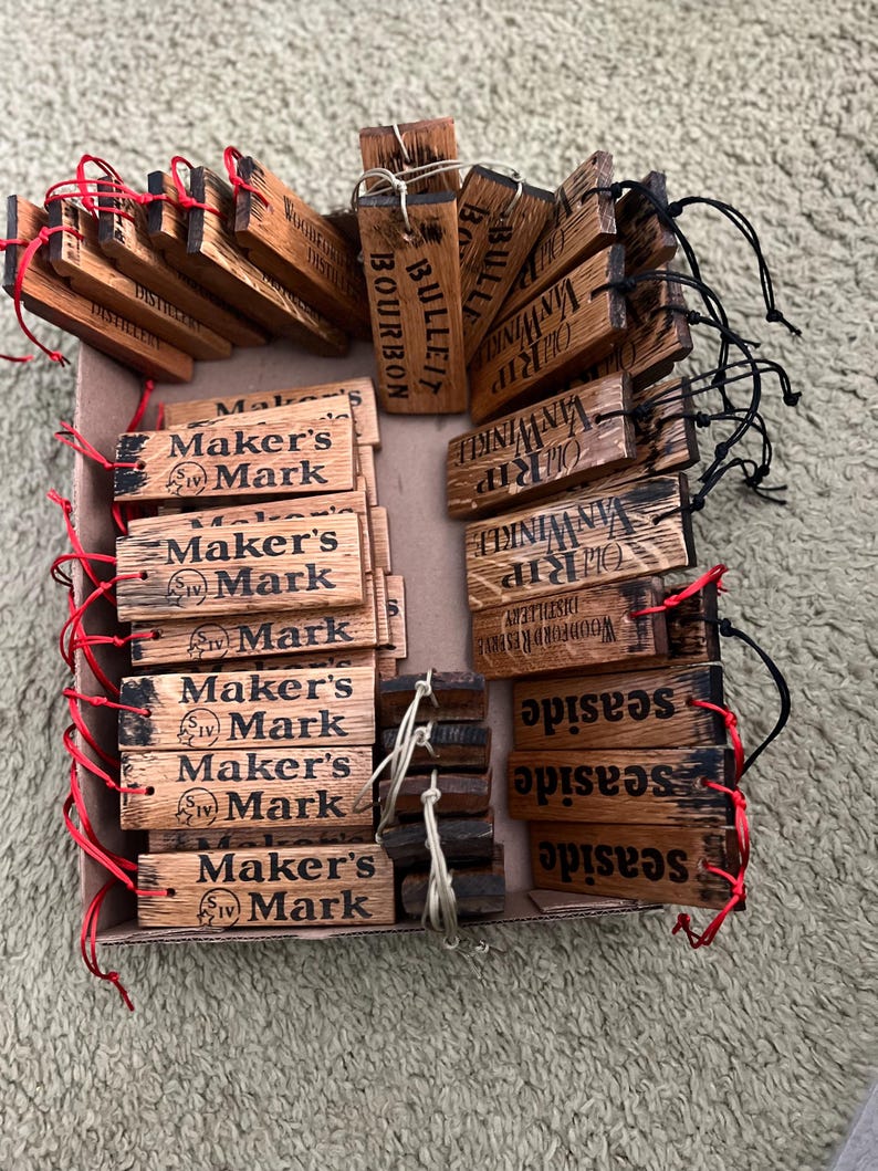 May include: A collection of wooden tags with various brand names and logos, including "Maker's Mark," "Old Rip Van Winkle," and "Woodford Reserve." The tags are rectangular, with dark text on a light brown wood grain. Some have red or black strings.