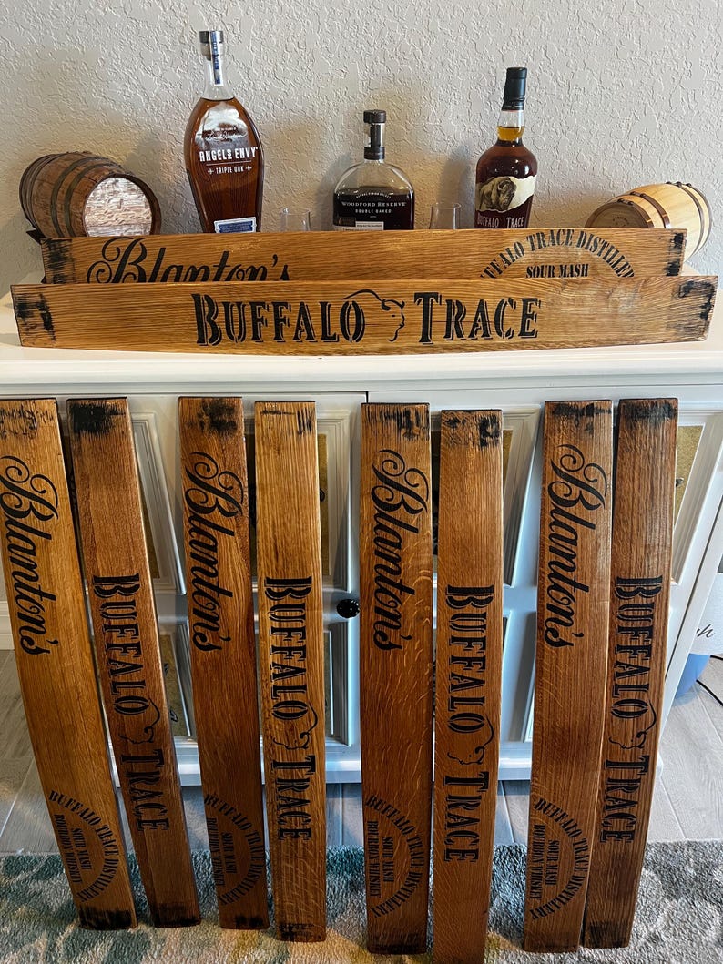 May include: Wooden barrel stave decor with the words "Blanton's" and "Buffalo Trace" in black lettering. The image also shows bottles of bourbon and small wooden barrels. The wood has a natural, aged appearance.