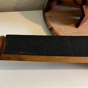 Puede incluir: Un elemento de madera rectangular con una superficie oscura y carbonizada y bordes de madera m&aacute;s claros. El elemento parece ser una secci&oacute;n de una duela de barril, mostrando la veta y la textura natural de la madera. La pieza es probablemente parte de un art&iacute;culo de decoraci&oacute;n r&uacute;stica.