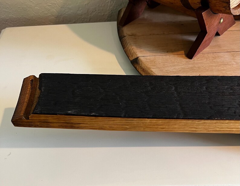 Puede incluir: Un elemento de madera rectangular con una superficie oscura y carbonizada y bordes de madera m&aacute;s claros. El elemento parece ser una secci&oacute;n de una duela de barril, mostrando la veta y la textura natural de la madera. La pieza es probablemente parte de un art&iacute;culo de decoraci&oacute;n r&uacute;stica.