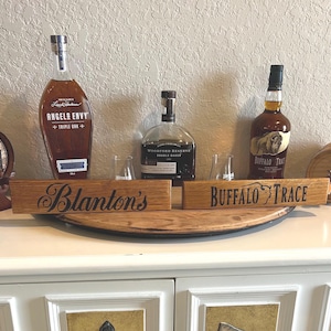 Puede incluir: Una exhibici&oacute;n de botellas de whisky y peque&ntilde;os barriles, incluyendo "Angel's Envy" y "Buffalo Trace." Dos letreros de madera dicen "Blanton's" y "Buffalo Trace." Las botellas contienen l&iacute;quido &aacute;mbar. La exhibici&oacute;n est&aacute; sobre una superficie blanca.