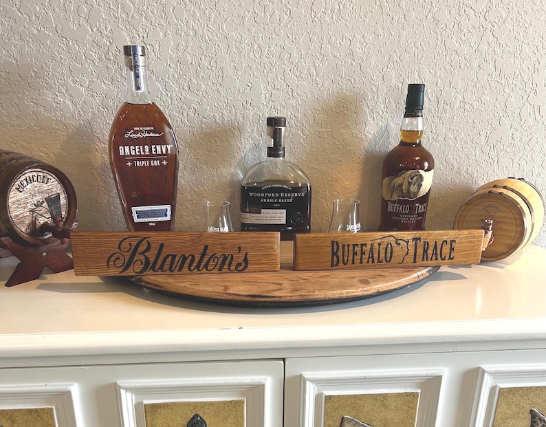 Puede incluir: Una exhibici&oacute;n de botellas de whisky y peque&ntilde;os barriles, incluyendo "Angel's Envy" y "Buffalo Trace." Dos letreros de madera dicen "Blanton's" y "Buffalo Trace." Las botellas contienen l&iacute;quido &aacute;mbar. La exhibici&oacute;n est&aacute; sobre una superficie blanca.