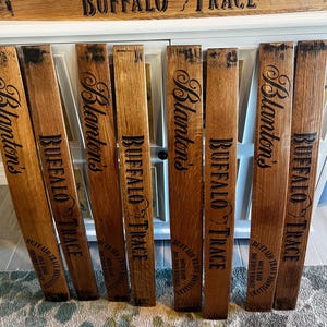 May include: Wooden barrel staves with the words "Blanton's" and "Buffalo Trace" printed in black. The staves are arranged in front of a white cabinet. The top of the cabinet has a wooden sign with the words "Buffalo Trace" printed on it.
