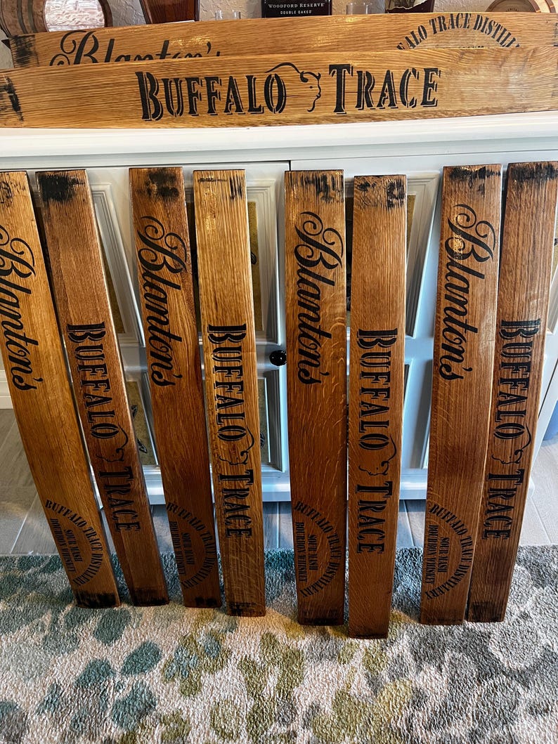 May include: Wooden barrel staves with the words "Blanton's" and "Buffalo Trace" printed in black. The staves are arranged in front of a white cabinet. The top of the cabinet has a wooden sign with the words "Buffalo Trace" printed on it.