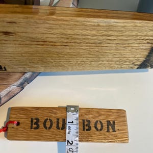 Puede incluir: Una etiqueta rectangular de madera con la palabra "BOURBON" impresa en negro. La etiqueta mide aproximadamente 2,5 cm de ancho. Una cuerda roja est&aacute; adjunta. La etiqueta es de madera marr&oacute;n claro.